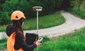 A female land surveyor using GPS equipment to perform a boundary survey on open, undeveloped land near a rural road